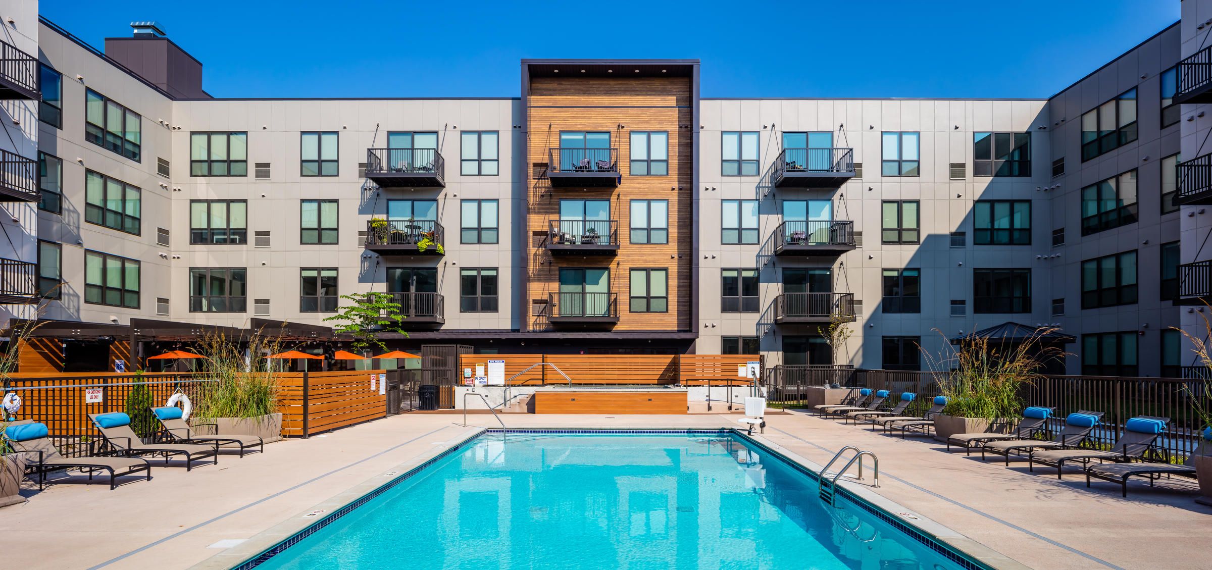 Modern apartment complex with balconies overlooking a swimming pool and lounge chairs on a sunny day.