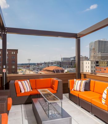 Modern rooftop patio with orange sofas, a fire pit table, and city buildings in the background under a blue sky.